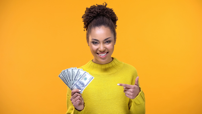 Smiling young lady pointing at dollar banknotes in hand, bank credit, earnings